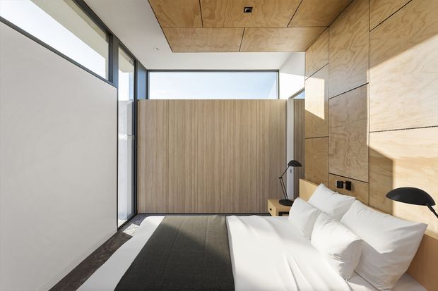 A compact, contemporary bedroom with a neatly made bed, white bedding, dark rug, timber veneer wall and ceiling panels, and matching vertical timber screening. Full-height glazing brings in strong daylight, while black bedside lamps and dark window frames provide contrast.