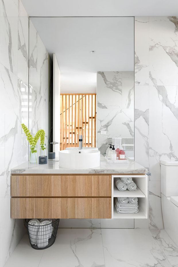Bright contemporary bathroom featuring full-height white marble-look wall and floor tiles with bold grey veining, a floating natural timber vanity with integrated open towel shelving, white vessel basin, frameless mirror, and a white toilet. A warm timber vertical slat screen is visible beyond the bathroom, adding contrast to the clean white scheme.