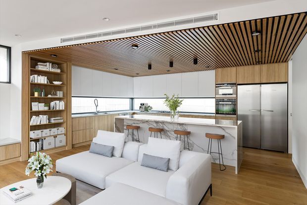 Open-plan contemporary kitchen and living area with a white sectional sofa in the foreground, a marble-look island with bar stools, light timber cabinetry and flooring, integrated stainless-steel appliances, open shelving, and a distinctive timber slatted ceiling. The bright interior has clean lines and a warm neutral palette.