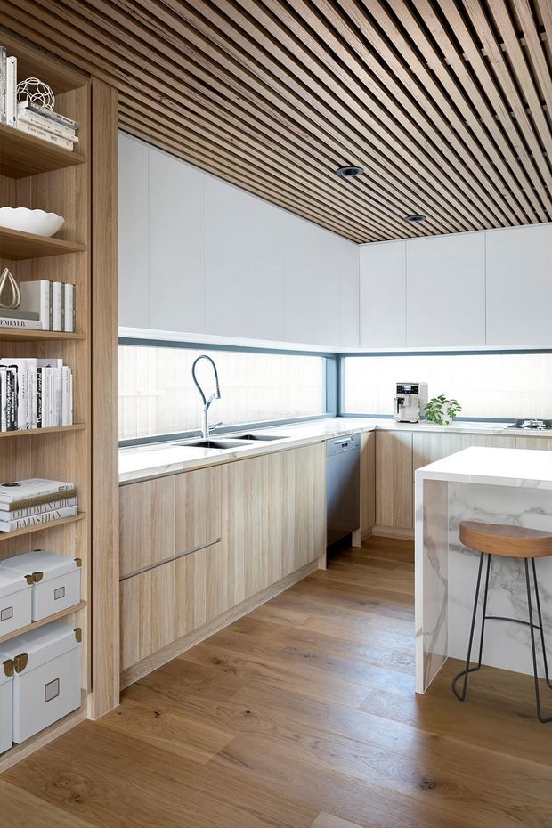 Bright contemporary kitchen with an L-shaped run of light timber cabinetry, white upper cabinets, expansive corner windows, a marble-look island, stainless-steel sink and appliances, and integrated open shelving. The ceiling features a distinctive slatted timber finish with recessed downlights, while the timber flooring creates a cohesive warm palette.
