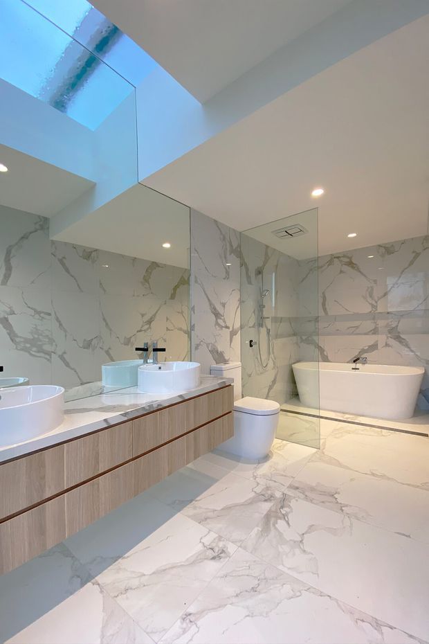 A bright contemporary bathroom with large-format white marble-look surfaces featuring grey veining on the walls and floor. The room includes a long floating timber vanity with multiple white vessel basins, black tapware, a wall-to-wall mirror, a glass-enclosed shower, a freestanding white bathtub, and a concealed toilet area. A large skylight and recessed ceiling lights provide abundant illumination.