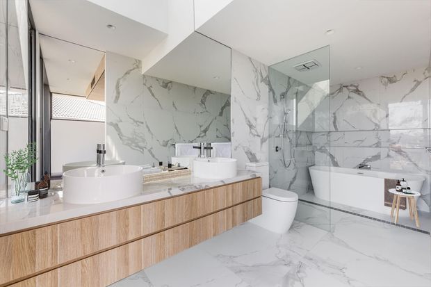 Bright, spacious contemporary bathroom with a floating natural timber double vanity, white vessel basins, wall-mounted mirrors, and extensive white marble-look veined tile surfaces. A frameless glass shower and freestanding white bathtub occupy the rear of the room, with a separate toilet and minimal chrome fixtures. Large glazing at the left brings in daylight and overlooks an exterior tiled roof.