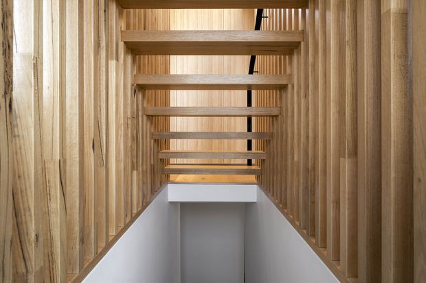 A narrow stairway or landing framed by extensive vertical and horizontal timber elements. Warm natural wood wall panelling and exposed stair structure create a rhythmic, tunnel-like architectural composition, with white-painted lower walls and a dark stair opening visible beyond.