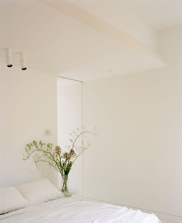 A serene, minimalist bedroom photographed from beside the bed. The room has smooth warm-white walls and ceiling, a simple white bed with textured white bedding and pillows, a small glass vase of delicate branches, and discreet recessed and surface-mounted spot lighting. A full-height pale panel or wardrobe door is visible at the rear, creating a clean, uncluttered composition.