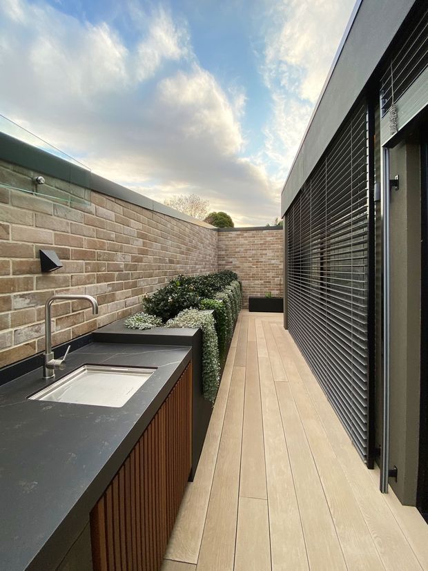 A narrow contemporary outdoor courtyard or service terrace enclosed by pale brick walls. The space features a built-in outdoor kitchen or utility counter with a dark benchtop, integrated sink and vertical timber-slat cabinetry, alongside raised planters with clipped greenery. Pale timber-look decking runs beside a dark louvred exterior wall and glazed door.