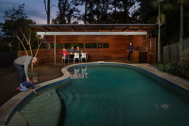 Twilight view of a landscaped residential swimming pool beside a timber-clad outdoor entertaining pavilion. The pool dominates the foreground, with a curved shallow ledge, dark decking, a long timber wall, outdoor dining setting, and a flat projecting pergola roof with exposed framing. Dense trees and planting surround the space, creating a private, integrated backyard setting.