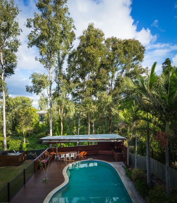 A lush tropical landscaped backyard viewed from above, featuring a curved swimming pool surrounded by timber decking, a compact covered outdoor entertaining pavilion with built-in seating and a dining area, and dense mature trees and palms. The pavilion has a low-pitched corrugated roof and dark timber finishes. Bright daylight and blue sky create a relaxed resort-like setting.
