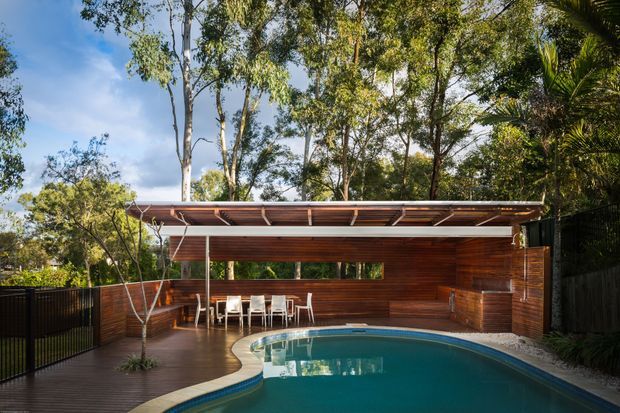 A landscaped outdoor pool area beside a compact open-air pavilion. The pavilion has warm timber wall cladding, a broad flat roof with exposed timber soffit and slender supports, an outdoor dining setting, and a built-in timber bar or counter. A curved swimming pool occupies the foreground, with timber decking, dark fencing, and dense mature vegetation surrounding the space.