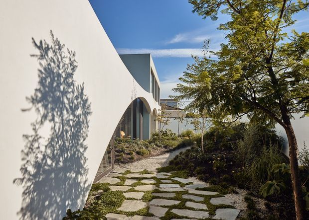 A contemporary curved white building exterior viewed along a landscaped garden path. The facade features smooth white walls, tall dark-framed glazing, arched openings, and a flat roofline. Irregular pale stepping stones set into gravel and low groundcover lead through dense planting and mature trees under a clear blue sky.