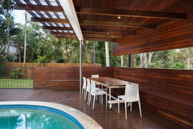 Covered outdoor dining and entertaining area beside a swimming pool, with a timber deck, dark horizontal timber wall cladding, timber-lined ceiling, exposed roof beams, and a slatted pergola. A long dining table with white chairs sits against the enclosed timber balustrade, surrounded by lush vegetation visible beyond the deck.