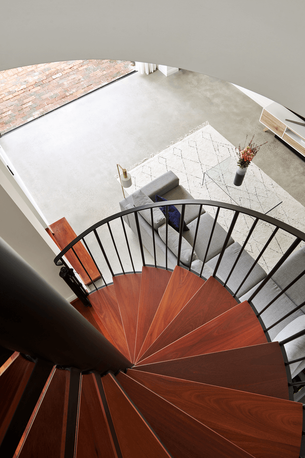 A high-angle interior view down a sculptural spiral staircase with rich reddish-brown timber treads, a dark central post, and slender black metal balustrades. Below is an open-plan living area with a grey sofa, pale patterned rug, concrete-look flooring, a low timber media console, and a vase of dried branches. A large opening at left reveals exposed brick paving or an adjacent brick surface, while soft neutral finishes create a bright, contemporary atmosphere.