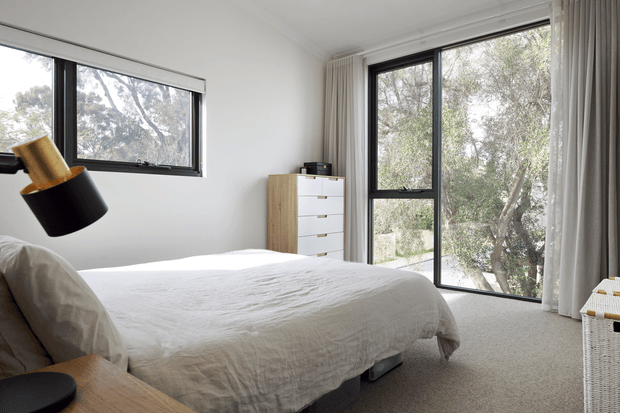 Bright, calm bedroom with a low-profile bed dressed in white textured bedding, pale timber bedside surface, light timber chest of drawers, and expansive black-framed windows overlooking leafy trees. Full-height curtains and neutral carpet create a soft, understated interior.