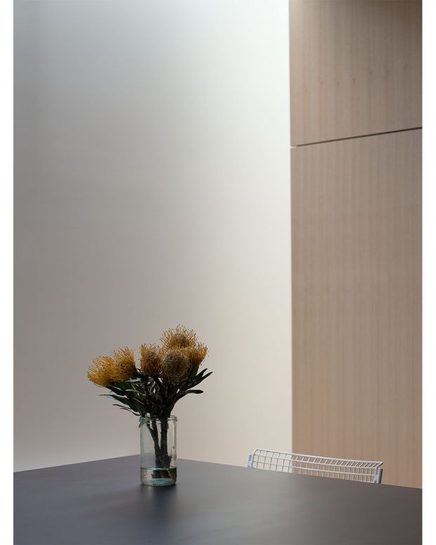 A minimalist dining area with a dark rectangular dining table, a clear glass vase containing dried botanical stems, and a partially visible wire-frame chair. The space has a plain warm-white wall and tall light timber built-in cabinetry or wall panelling, creating a restrained contemporary interior with soft natural light.