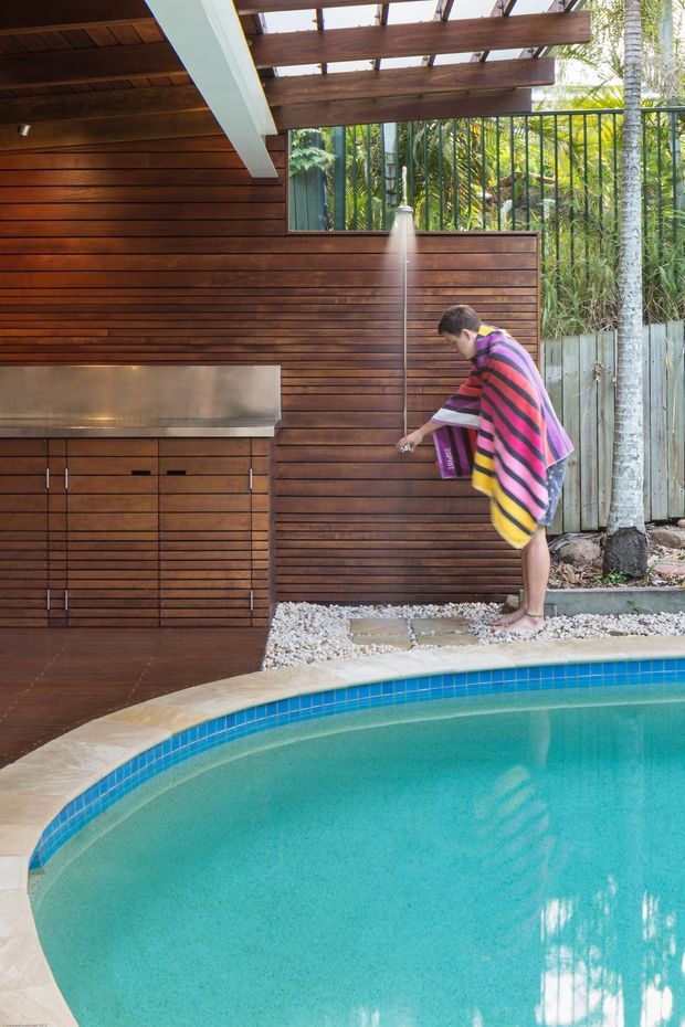 A tropical outdoor pool area with a curved swimming pool in the foreground and a timber-clad outdoor kitchen or barbecue enclosure behind it. The space includes a stainless-steel built-in barbecue, outdoor shower, pergola with exposed timber beams, slatted timber screening, pebble landscaping, and lush planting beyond a metal fence. A person in a striped towel is using the shower.