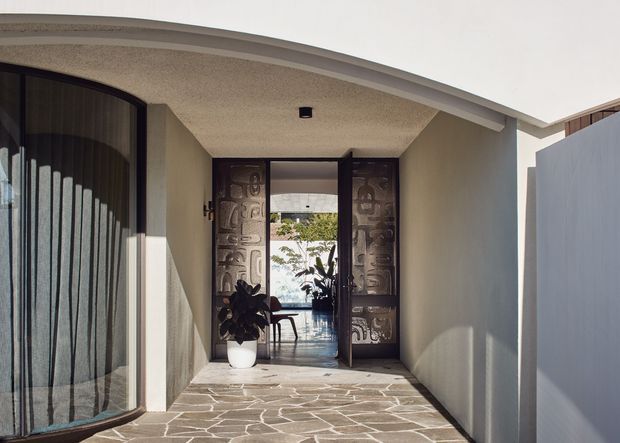 A covered residential entryway with a curved roof overhang, textured light walls, dark timber-framed glazing, and an open decorative glass entry door. Irregular natural-stone paving leads through the vestibule toward a bright interior and a glimpse of greenery beyond. The scene has a refined mid-century-influenced character with strong indoor-outdoor connection.