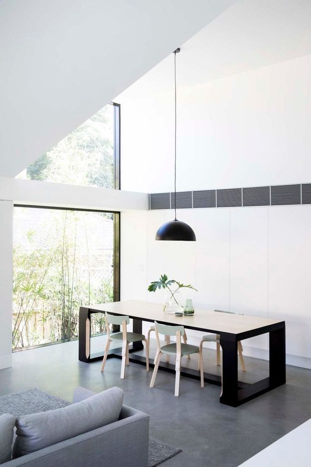 Bright minimalist open-plan interior featuring a dining area beside floor-to-ceiling glazing. A long light-toned dining table with dark structural legs is paired with pale wooden chairs, beneath a suspended black dome pendant. White walls and cabinetry, polished grey concrete flooring, a grey sofa, and abundant natural light create a clean contemporary atmosphere. A high-level linear ventilation grille and angled ceiling plane add architectural definition.