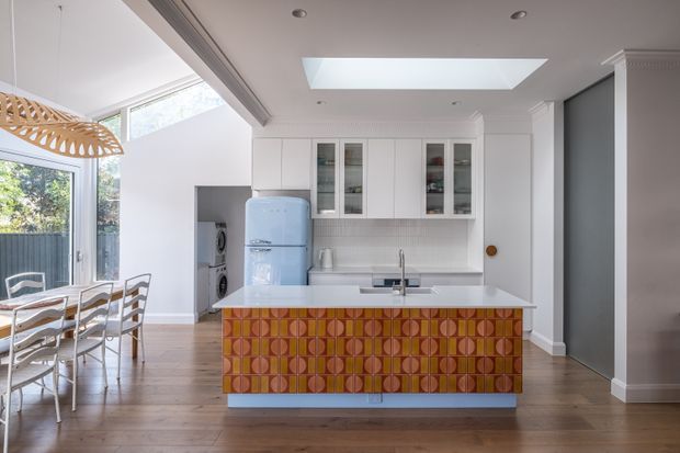 Bright open-plan kitchen and dining area with white cabinetry, a pale blue retro-style refrigerator, timber flooring, skylight, and large glazed openings. The kitchen island has a bold patterned orange, terracotta, and mustard geometric tile feature, while a sculptural woven pendant hangs above the dining table.