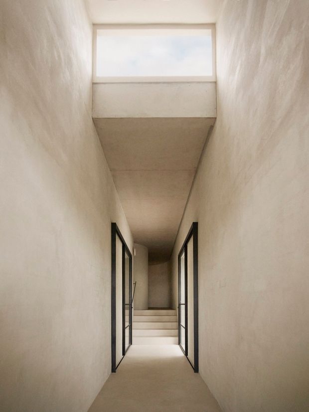 A narrow, minimalist interior hallway with warm off-white textured plaster walls and ceiling, pale flooring, and black-framed glazed doors opening along both sides. A short flight of pale steps leads toward the back, while a high rectangular clerestory window brings in soft daylight. The composition is quiet, linear, and monolithic.