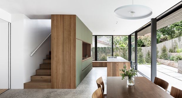 Bright contemporary open-plan dining and kitchen area with a timber dining table in the foreground, a built-in timber and muted green cabinetry volume enclosing a staircase, and a kitchen beyond. Full-height black-framed glazing opens the room toward a landscaped courtyard, while a large circular pendant hangs from the white ceiling. The space combines warm timber, terrazzo-style flooring, clean white surfaces, and indoor-outdoor connection.
