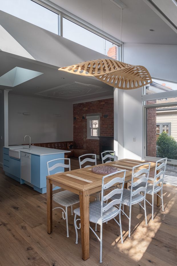 Bright open-plan interior combining a kitchen and dining area, with a timber dining table and white metal chairs in the foreground, a pale blue island kitchen to the left, exposed red-brick walls, timber flooring, large glazed openings, and a sculptural suspended timber pendant light. A lounge area is visible beyond the kitchen, creating a spacious, loft-like atmosphere.