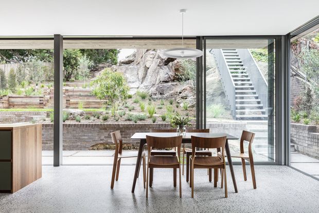 Bright contemporary dining area with a timber dining table and six timber chairs, positioned beside extensive floor-to-ceiling glazing. The room opens visually onto a landscaped terraced garden with exposed rock, brick retaining walls, and an external stairway. A partial timber kitchen cabinet is visible at left, while a slim pendant light hangs above the table. The space has a restrained material palette and strong indoor-outdoor connection.