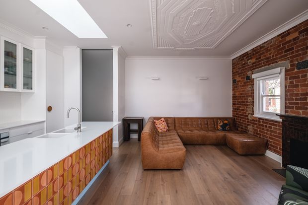 Open-plan kitchen and living room with a white island featuring colourful patterned tile cladding, white cabinetry, timber flooring, a cognac-brown sectional sofa, exposed red brick walls, a black fireplace, and an ornate pressed ceiling with a large skylight. The space blends contemporary kitchen fittings with traditional architectural detailing and warm eclectic furnishings.