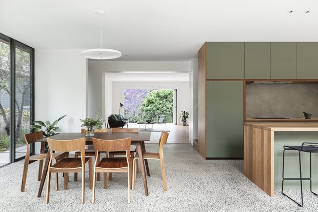 Open-plan contemporary kitchen and dining area with a timber dining table and chairs, terrazzo flooring, pale walls, extensive glazing, and muted green built-in cabinetry. A compact kitchen with a wood-fronted island and integrated concrete-look splashback opens toward a bright living area and garden view.