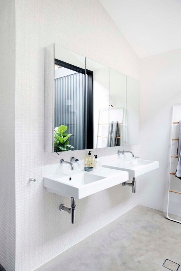 Bright minimalist bathroom with two wall-mounted white rectangular basins, exposed chrome plumbing and wall-mounted chrome taps beneath a large mirrored cabinet. The walls are clad in small white mosaic tiles, while the floor has a smooth light-grey concrete-like finish. A glass shower enclosure with dark framing and a timber towel ladder are visible in the mirror and at the right edge.