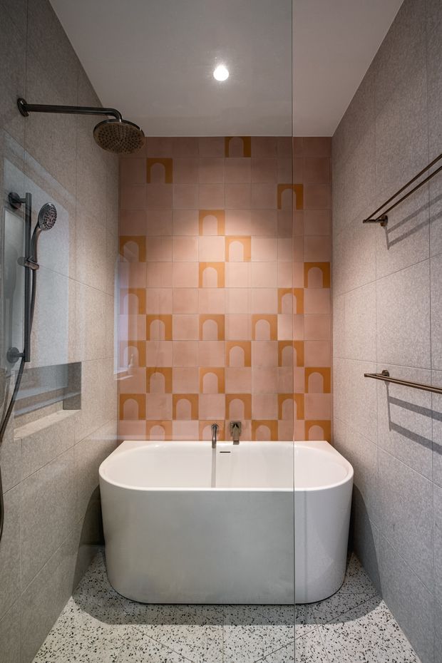 A compact contemporary bathroom featuring a freestanding white bathtub enclosed by a clear glass shower screen. The shower has dark metal fixtures and a rainfall head, while the walls are clad in pale grey textured tiles. A terracotta and peach patterned tile feature wall with repeated arch motifs adds a warm graphic accent. The floor is finished in light terrazzo with dark aggregate, and simple dark metal towel rails are mounted on the right wall.
