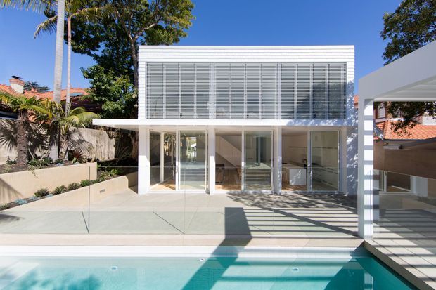 Bright exterior view of a contemporary two-storey residence beside a swimming pool. The white cubic building has extensive glazing, a screened upper level with horizontal louvres, a covered ground-floor terrace, and a separate white carport structure at right. Strong geometric shadows fall across the paved pool terrace and water.