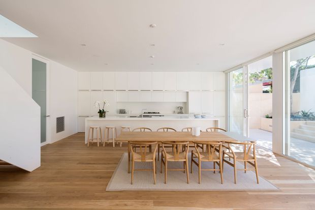 Bright open-plan kitchen and dining area with extensive white built-in cabinetry, a long island with stools, and a large timber dining table surrounded by woven timber chairs. Warm timber flooring, white walls and cabinetry, recessed lighting, and full-height glazed sliding doors create a light, minimalist space opening to a paved outdoor area. A skylight is visible at the upper left.
