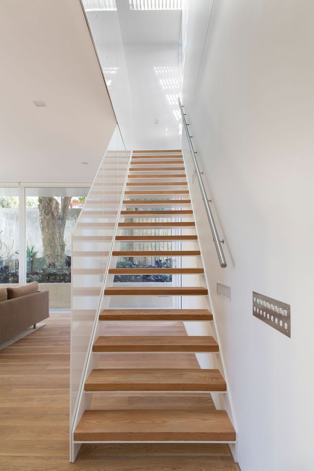 Bright contemporary stairway with open-riser timber treads, slim white-painted stringers, clear glass balustrade on one side, and a stainless-steel handrail on the other. The stair rises through a tall white-lined void with a skylight or high-level glazing above. Timber flooring continues into the adjacent living area, visible at left through the open stair space.