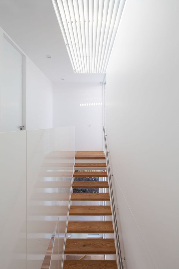 Bright minimalist interior view of a contemporary stairway ascending through a narrow white stair hall. The staircase has open-riser timber treads, slim metal handrails, and a clear glass balustrade. A large white linear slatted ceiling feature forms a strong architectural focal point, with daylight filtering through the open stair structure.