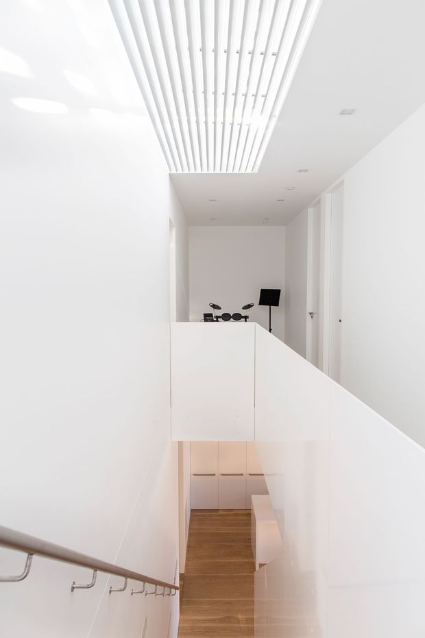 A bright minimalist interior stairwell and landing viewed from the stairs. White walls, balustrades and doors surround a narrow circulation space, with warm timber flooring below. A sculptural linear slatted ceiling feature brings daylight into the upper level, while a small black drum kit and floor lamp are visible at the landing.