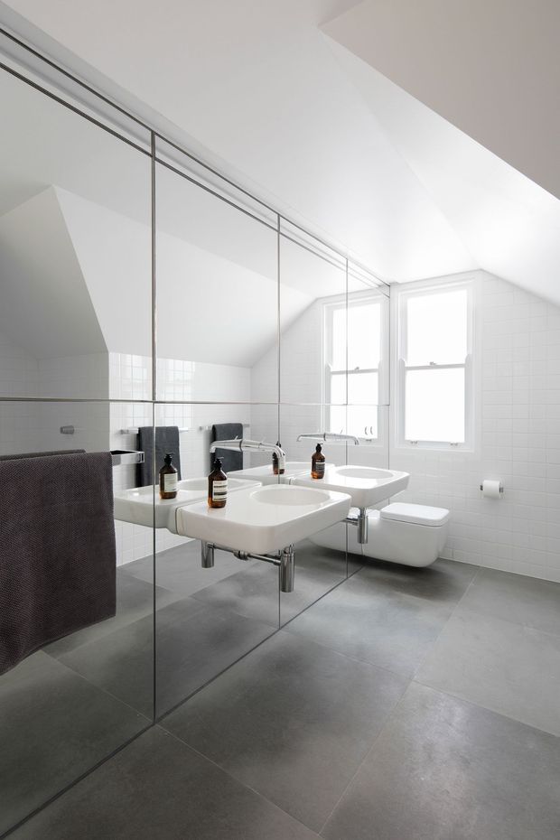 A bright, minimalist bathroom beneath a sloped ceiling, featuring two wall-mounted white ceramic basins with chrome taps, a wall-hung toilet, large grey floor tiles, white tiled walls, twin sash windows, and a full-height mirrored wall that reflects the fixtures. Dark towels and amber soap dispensers provide small accents.