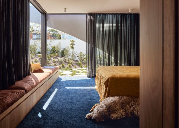 A contemporary bedroom with floor-to-ceiling glazing and dark sheer curtains opening onto a landscaped garden courtyard. The room features a low built-in timber window seat with dark cushions, a bed dressed in warm mustard bedding, a deep blue carpet, and a shaggy brown rug. Strong natural sunlight creates a bright, relaxed indoor-outdoor atmosphere.
