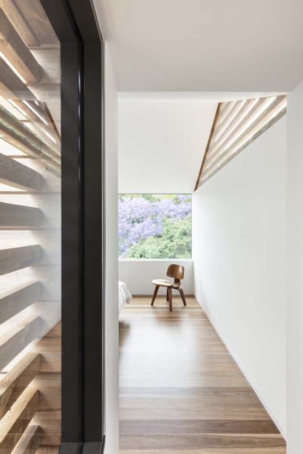 A narrow, minimalist interior hallway or landing with pale timber flooring, white walls and ceiling, and a black-framed glazed opening on the left. A compact timber chair sits at the end beside a large window overlooking foliage. Exposed timber slatted detailing is visible along the left and upper right, creating a warm architectural accent in an otherwise bright, restrained space.