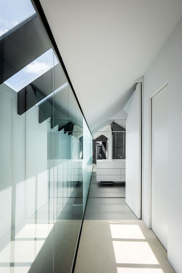 A bright, narrow minimalist interior corridor leading toward a compact bathroom vanity. One side is formed by a full-height glass partition reflecting the space and dark structural elements, while the opposite side has smooth white walls and flush doors. Large skylight-like openings cast rectangular sunlight across the pale floor. At the end, a white floating vanity, mirror, and dark tiled wall are visible.