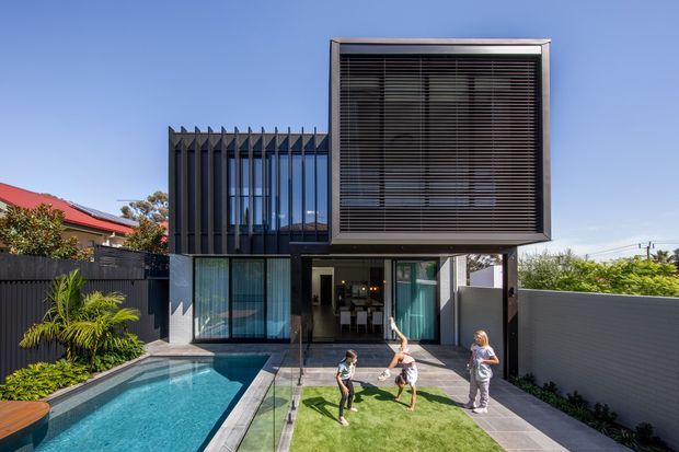 Exterior view of a contemporary multi-level residence with a striking projecting upper box clad in dark horizontal screening and extensive glazing. The home features a covered ground-floor entertaining area, swimming pool, paved terrace, and a small lawn courtyard, with neighbouring homes and landscaping visible around it.