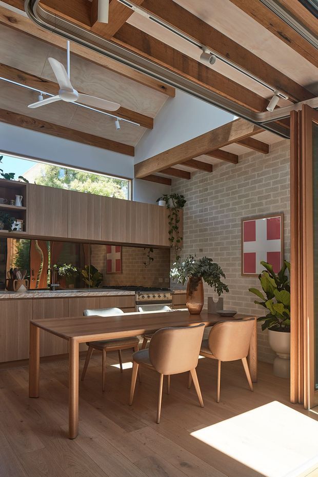 Warm open-plan dining and kitchen interior with a timber dining table and upholstered chairs in the foreground. The space features extensive timber cabinetry and flooring, exposed timber ceiling beams, exposed services and track lighting, a ceiling fan, pale painted walls, and a grey brick feature wall. Large windows and glazing bring in abundant natural light, while indoor plants and framed artwork add softness and colour.