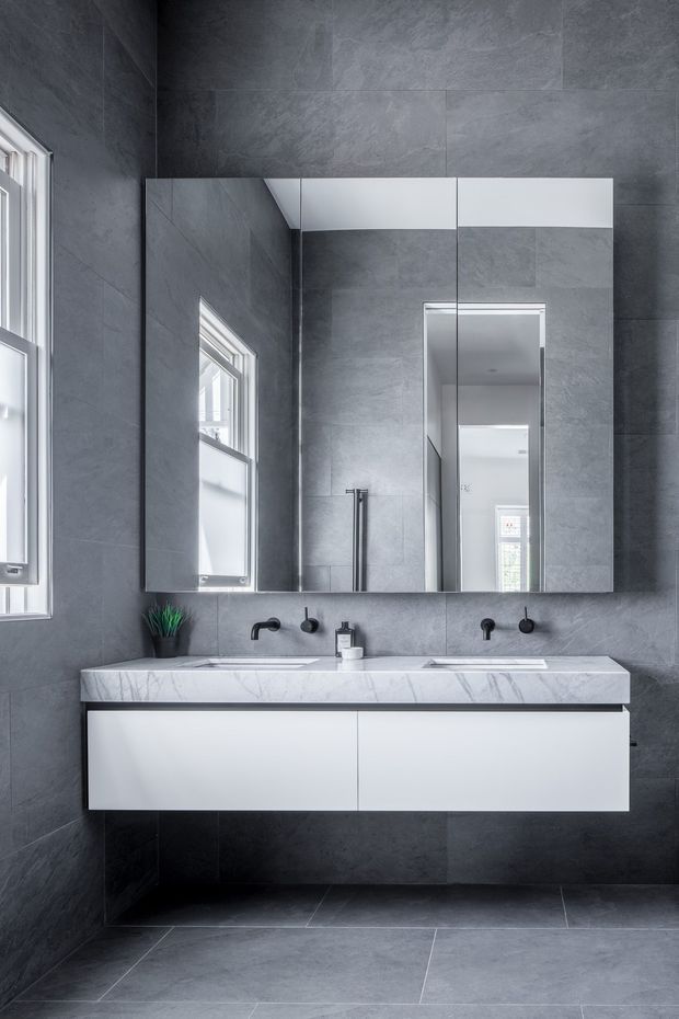 A contemporary bathroom with a double floating vanity, white cabinetry, a pale veined stone benchtop, twin black wall-mounted tap sets, mirrored storage above, and large dark grey stone-look wall and floor tiles. A window provides natural light, while a doorway reflection shows an adjoining space.