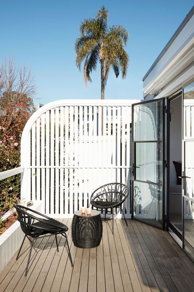 A bright outdoor terrace or balcony with grey-brown timber decking, black woven lounge chairs, and a small round side table. A white vertical slatted privacy screen with a curved top forms the backdrop, while black-framed glazed folding or hinged doors open into the adjacent interior. Palm trees and flowering vegetation are visible beyond the screen under a clear blue sky.