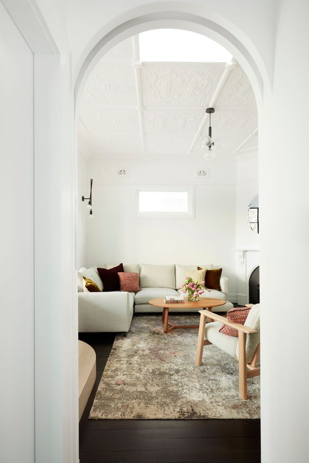 Bright, narrow living room viewed through a broad arched doorway. The room has white walls and ornate pressed ceiling panels, a white sectional sofa with colourful cushions, a round timber coffee table, a patterned area rug over dark timber flooring, a light timber-framed lounge chair, wall sconces, and a white fireplace surround. The space combines traditional architectural details with restrained contemporary furnishings.