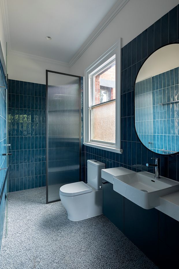 A compact, refined bathroom featuring deep blue vertical wall tiles, a terrazzo-style pebble floor, a wall-mounted white basin with dark fixed cabinetry, a modern white toilet, a black-framed translucent glass shower screen, and a large arched mirror. White decorative ceiling moulding and a frosted window provide contrast and natural light.