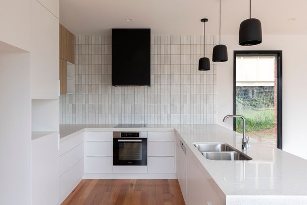 A compact contemporary kitchen with a U-shaped white built-in layout, integrated oven and cooktop, stainless-steel double sink, pale speckled benchtops, and a full-height grid-pattern tiled splashback. Warm timber flooring and timber cabinetry at the rear contrast with the white joinery. Black pendant lights and a black-framed window add bold accents, with greenery visible outside.