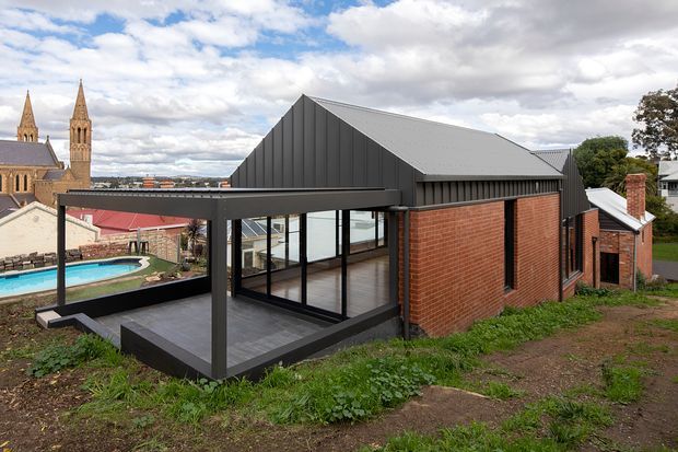 Exterior view of a contemporary extension with a steep gabled, dark metal-clad roof, red brick walls, extensive black-framed glazing, and a covered outdoor deck. The building sits on a sloping site beside a swimming pool, with historic stone church towers visible in the background.