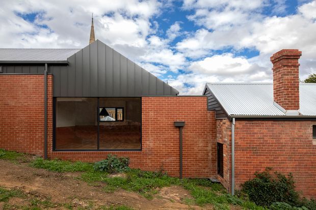 Exterior view of a red-brick building complex with a contemporary dark metal-clad gabled extension, corrugated metal roofs, large glazed opening, and a prominent traditional brick chimney. A slender church-like spire rises behind the roofline, with a partly cloudy sky and landscaped earth foreground.