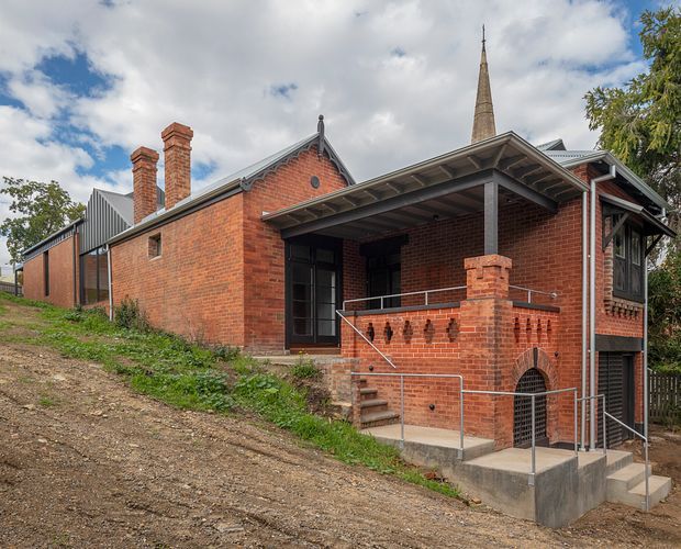 Exterior view of a historic red-brick building on a sloping site, featuring a steep gabled roof, twin brick chimneys, a prominent narrow spire, covered entry porch, brick balustrade, and contemporary metal accessibility railings. Mature trees and a partly cloudy sky surround the building.