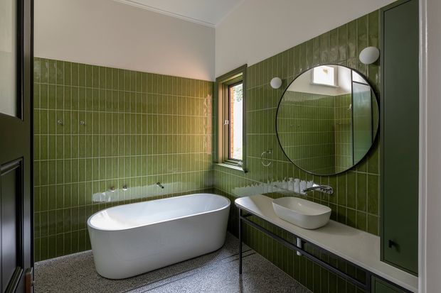 A compact contemporary bathroom with a freestanding white bathtub, wall-mounted basin on a dark vanity, round black-framed mirror, and green vertically grooved wall tiles. The floor is finished in terrazzo, with dark green built-in cabinetry and a narrow window providing natural light.