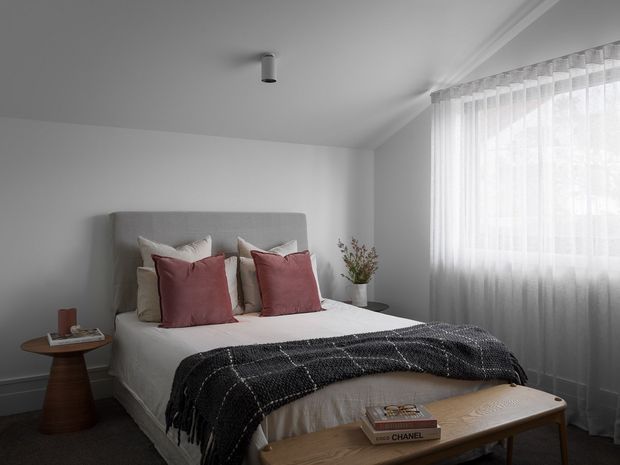 A softly lit, minimalist bedroom featuring a neatly made bed with a tall upholstered grey headboard, layered white and blush cushions, and a dark charcoal throw. A warm timber bedside table and low timber bench add contrast, while sheer floor-to-ceiling curtains diffuse daylight from the large window. The room has plain white walls, a simple ceiling-mounted light, and a calm contemporary styling.